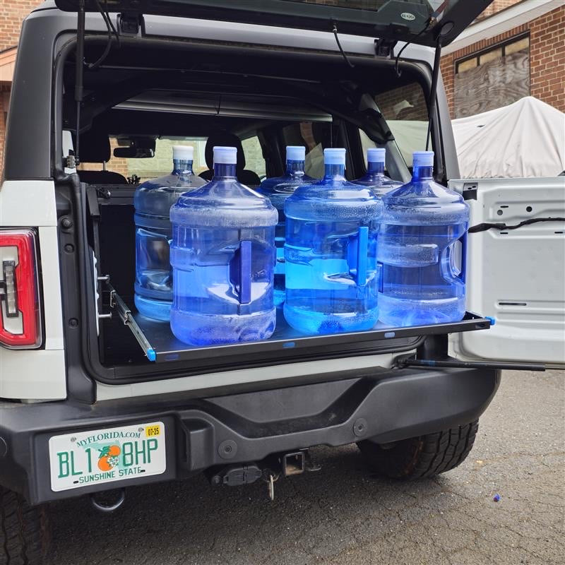 Closer angled view of the extended EZ4X4 Extreme EZTRUNK shelf on a white Ford Bronco supporting six full five-gallon water jugs.