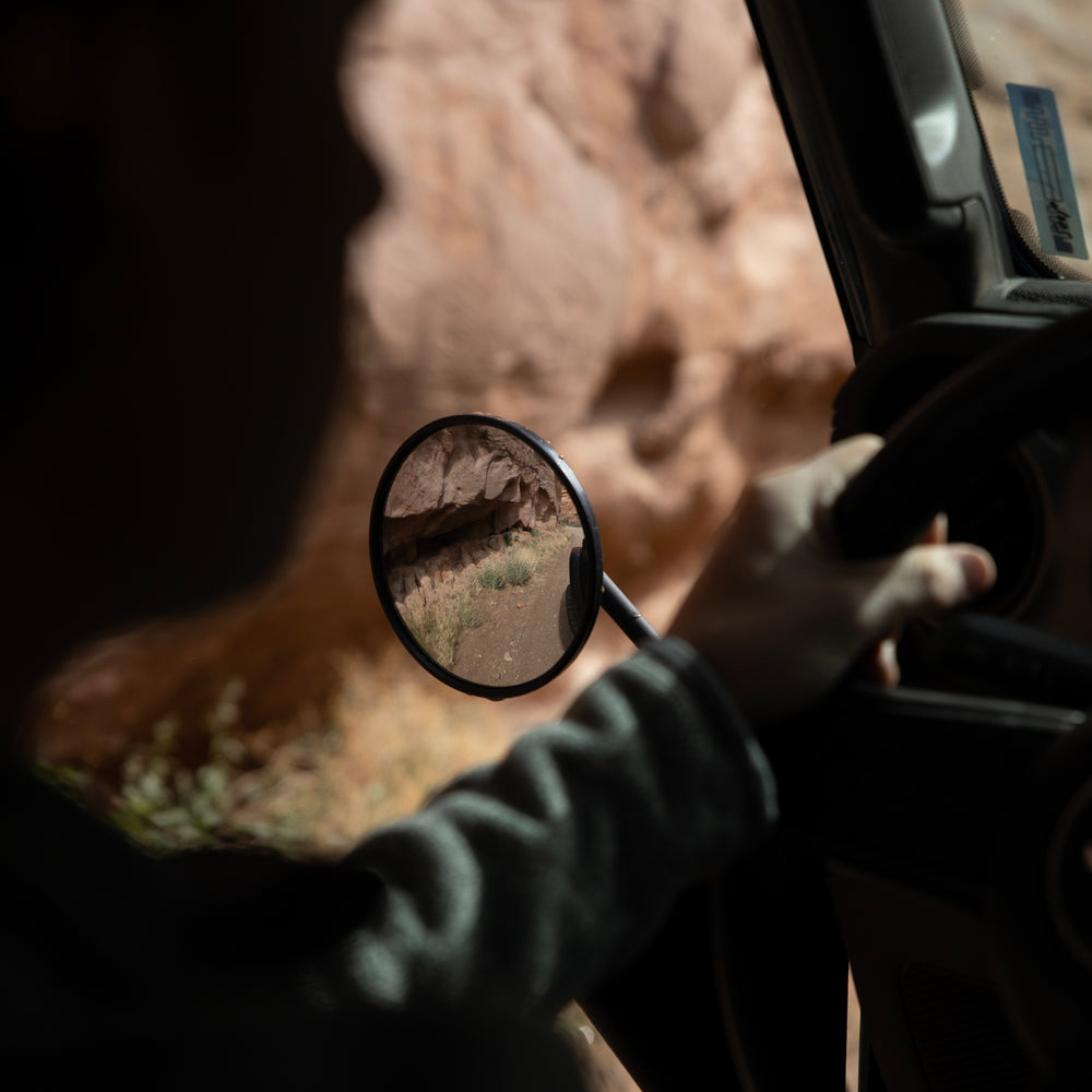 Person adjusting round aluminum Jeep mirror with desert reflection, showing off-road style and rugged visibility.