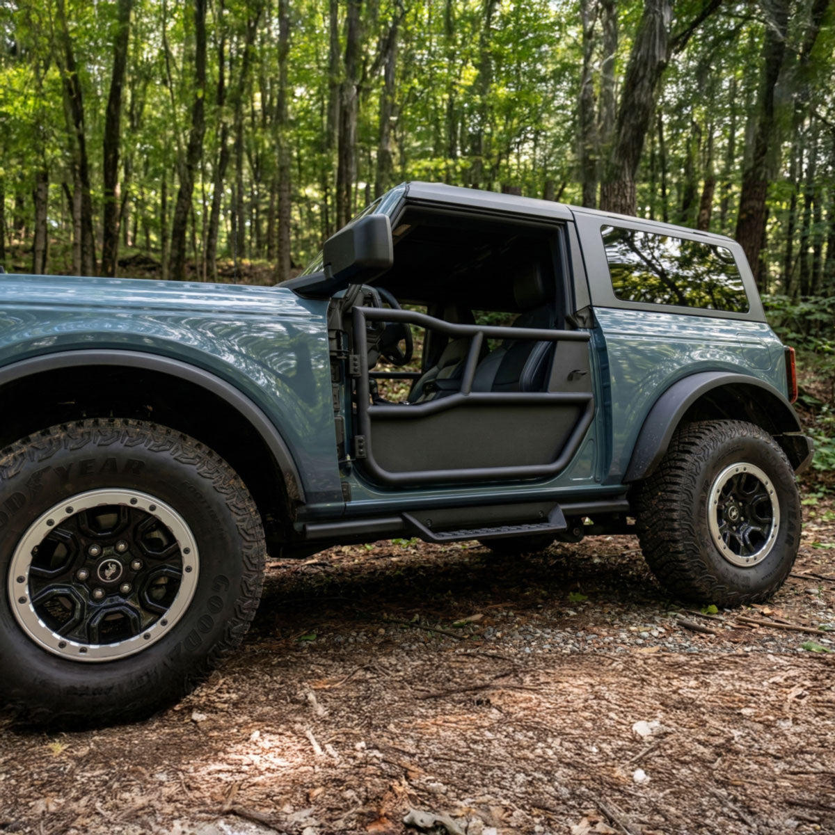 Blue 2-door Ford Bronco fitted with EZ4X4 Alpha Tube Doors and bottom All-Weather Panel set, parked on a wooded trail.