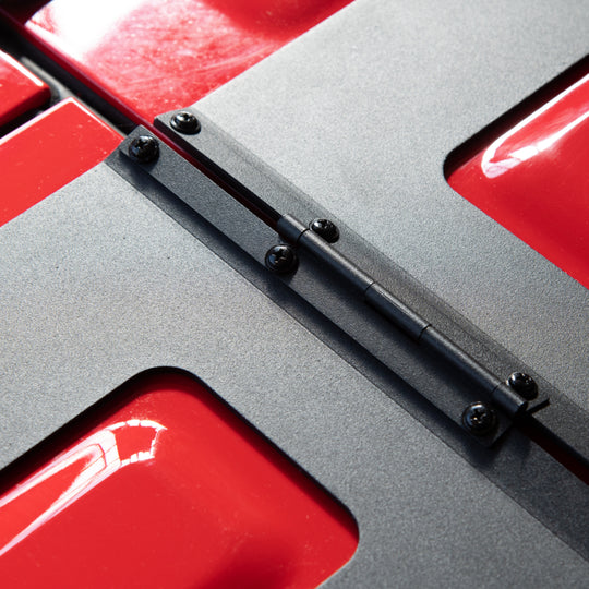 Close-up of the EZ4X4 EZ FlipTop hinge system mounted on a red Jeep, highlighting the black textured matte finish and heavy-duty bolt-on hardware at the hinge mounting point.