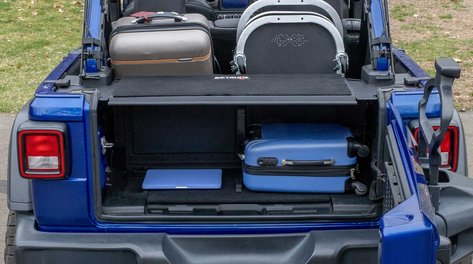 EZ4X4 EZTRUNK installed in a blue Jeep Wrangler, showing a suitcase and laptop stored inside the EZTRUNK with an additional suitcase and door cart positioned behind it.