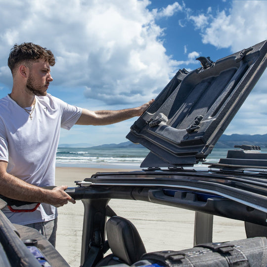 Man flipping black EZ Flip-Top panel on Jeep JT Gladiator at the beach, showing easy roof removal for open-air, off-road fun.