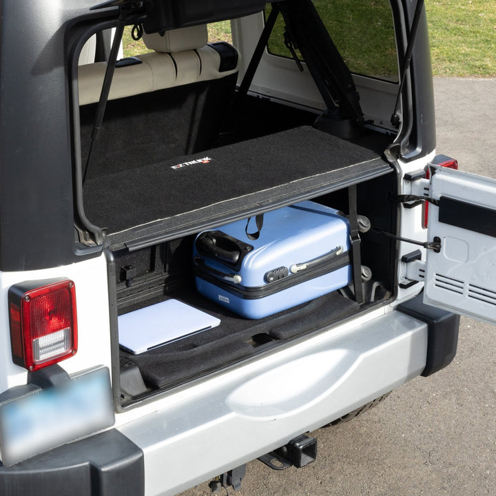 Black EZ4X4 EZTRUNK tailgate carpet installed in white 4-door Jeep Wrangler with suitcase and laptop stored inside.