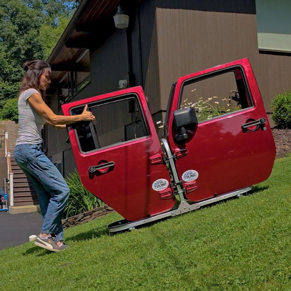 Woman rolling EZ4X4 aluminum door cart with four red Jeep doors down a hill, showing portable, durable storage design.