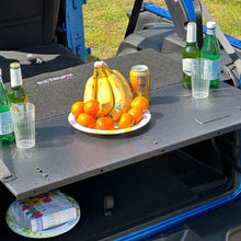 EZTrunk Tailgate Extension Table open in Jeep with fruit and drinks on top, plates and napkins stored on shelf below.