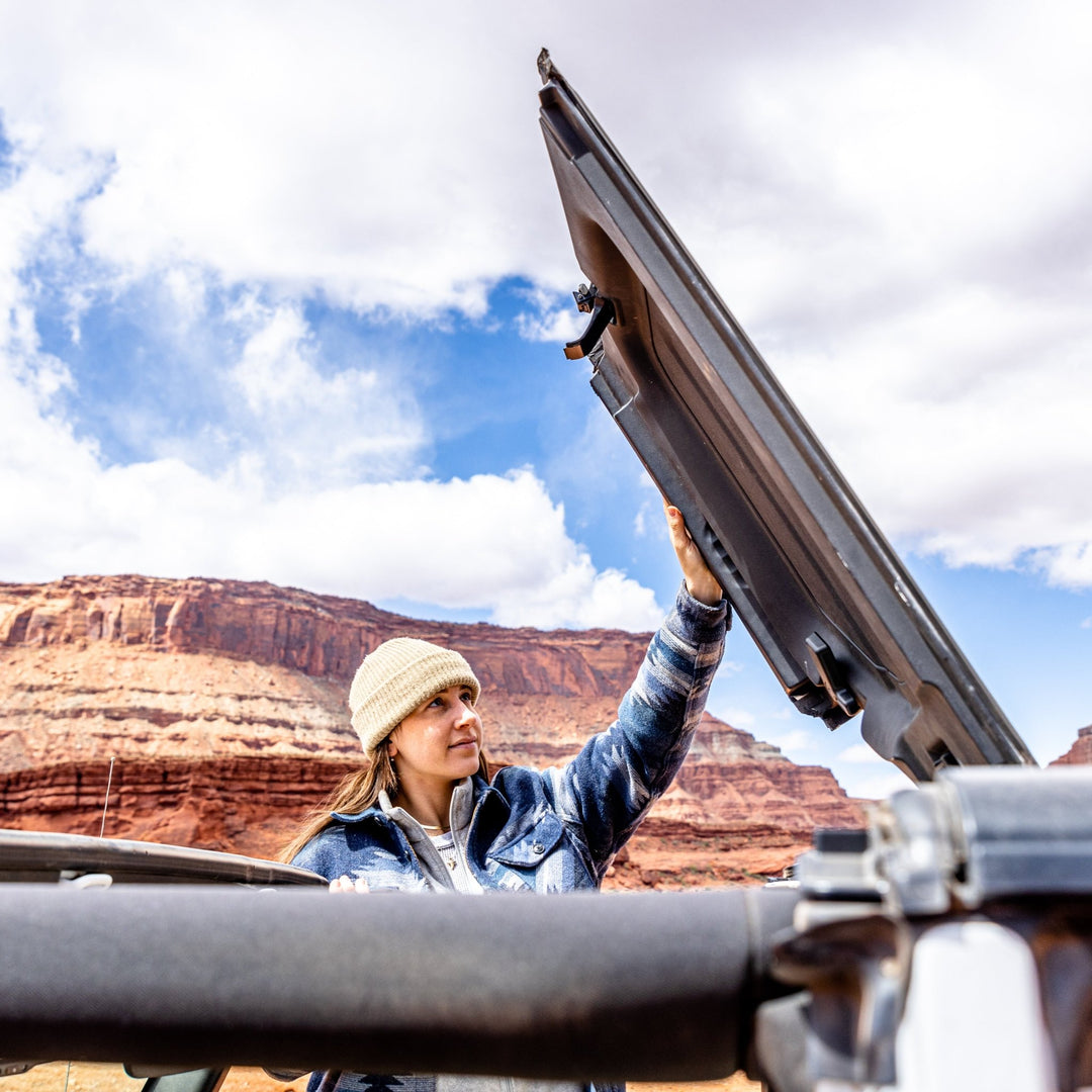 Woman flipping up black roof panel on Jeep Wrangler in desert, showcasing easy EZ FlipTop system for one-person operation.