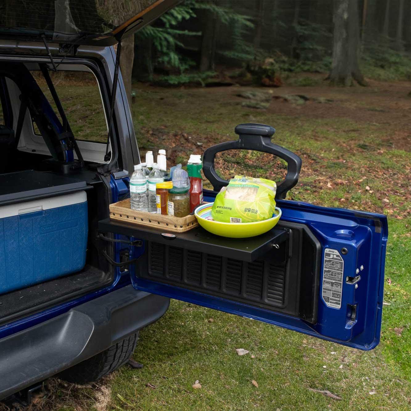 EZ4X4 Single Level Tailgate Table fully deployed on a blue Jeep Wrangler tailgate, holding a picnic setup of food and drinks in a forest setting.