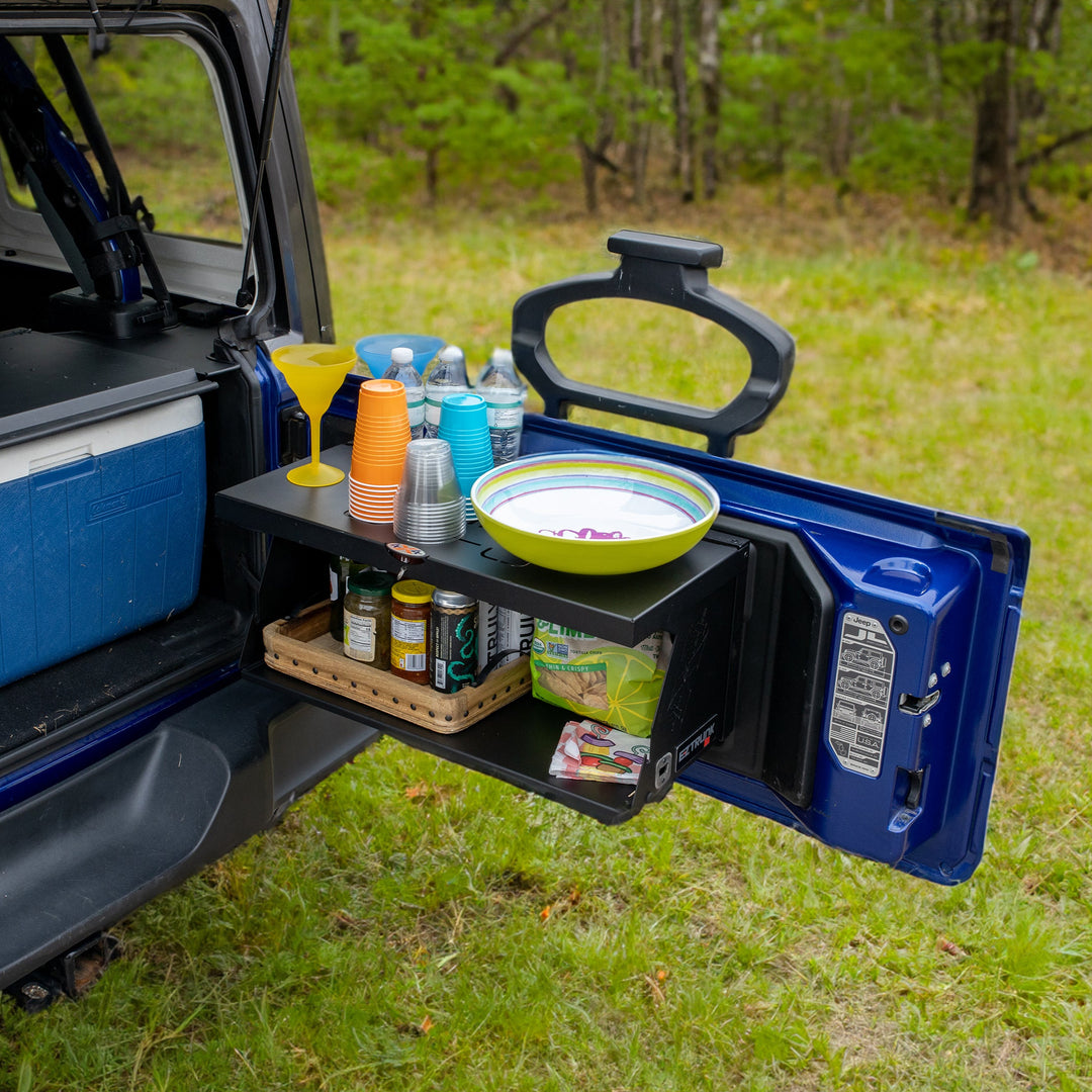 Angled view of black foldable tailgate table mounted on Jeep, shown holding food and drinks—perfect for camping and tailgating.