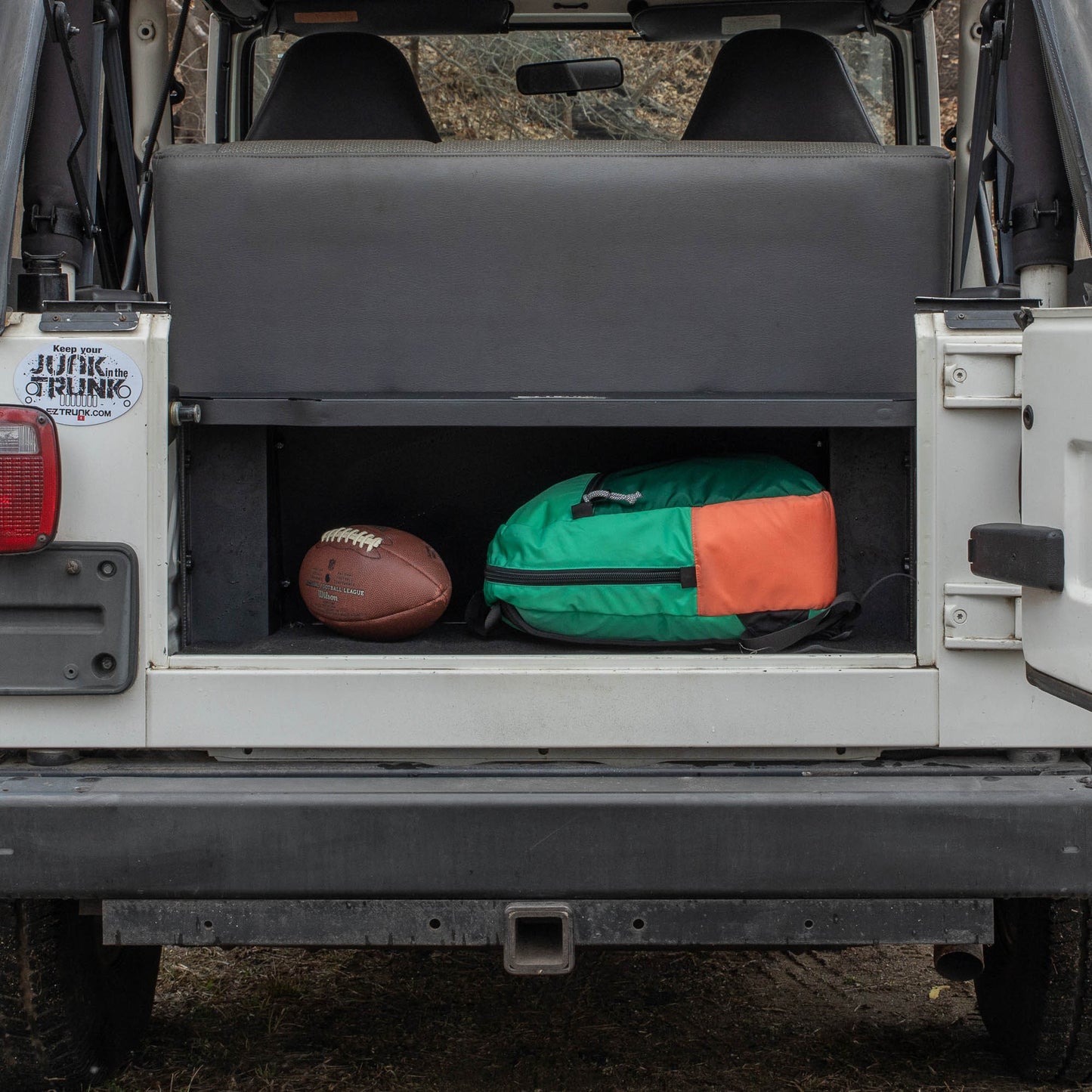 Black EZTrunk installed in cargo area of white Jeep TJ Wrangler with trunk door open, securing a backpack and football.