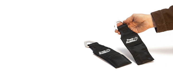Two black Truck Buddiez bed anchor straps with metal carabiners, one held by a hand.