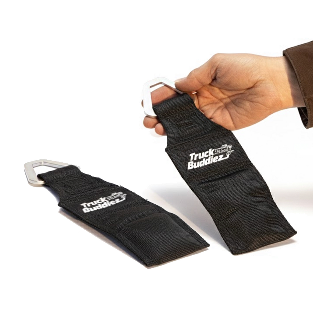 Two black Truck Buddiez bed anchor straps with metal carabiners, one held by a hand.